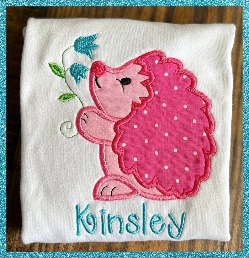 May include: A white onesie with a pink hedgehog with white polka dots and blue flowers embroidered on it. The name "Kinsley" is embroidered in teal below the hedgehog.