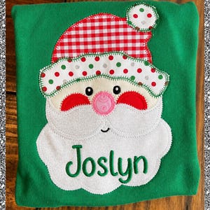 May include: A green shirt featuring a fabric applique of a Santa Claus face. The Santa has a red and white gingham hat, a white brim with red and green polka dots, a pink nose, and a white beard with the name Joslyn embroidered in green.