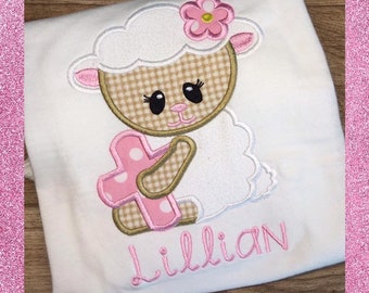 May include: White embroidered t-shirt with a pink and brown lamb holding a pink cross. The lamb has a pink flower on its head. The name "Lillian" is embroidered in pink below the lamb.