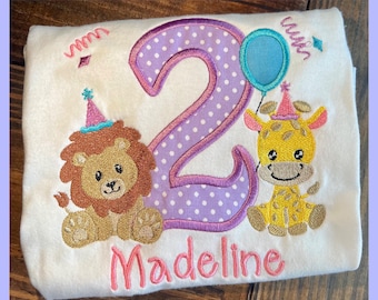 May include: White t-shirt with an embroidered design for a second birthday. Features a large purple number 2 with white polka dots, a lion, a giraffe, a balloon, and the name "Madeline" in pink.