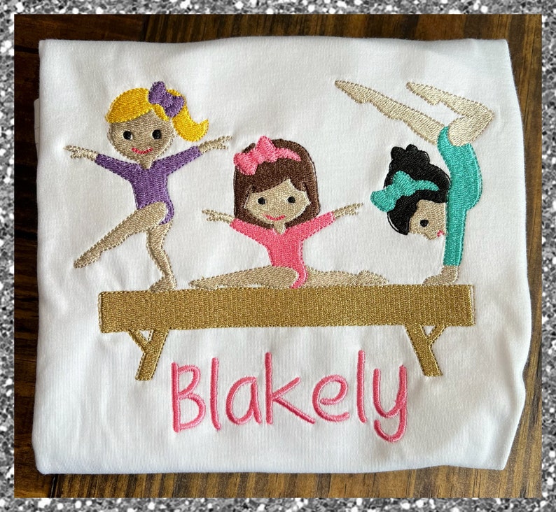 Op de afbeelding: Een wit T-shirt met een geborduurd ontwerp van drie meisjes die turnen op een evenwichtsbalk. De meisjes dragen verschillende gekleurde turnpakjes en hebben verschillende gekleurde haren. De tekst "Blakely" is onder de balk geborduurd.