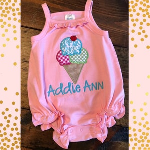 May include: Pink baby romper with spaghetti straps and ruffle trim. Features an ice cream cone design with three scoops in blue, pink, and green, topped with a cherry. The name "Addie Ann" is embroidered in teal below the ice cream.