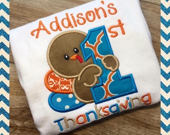 May include: A white onesie with an embroidered turkey holding a blue number one and the text "Addison's 1st Thanksgiving".