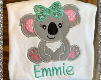 Könnte beinhalten: Ein weißes T-Shirt mit einem grauen Koalabären, der eine grüne Schleife mit Glitzer trägt. Der Koala sitzt mit rosa Pfoten. Der Name "Emme" ist in Grün unter dem Koala gestickt.