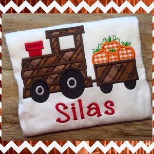May include: A white t-shirt with a brown and plaid train pulling a wagon full of orange pumpkins. The wagon and train have black wheels. The name Silas is embroidered in red on the shirt.