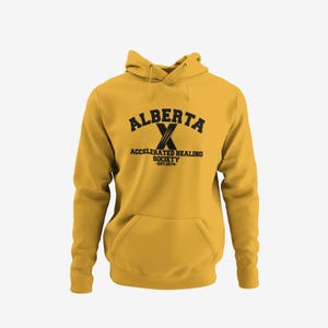 May include: Mustard yellow hoodie with a front pocket and drawstring hood. The front features the text "ALBERTA" above a large "X" and the words "ACCELERATED HEALING SOCIETY" and "EST.1874" in black lettering. A casual, comfortable garment.