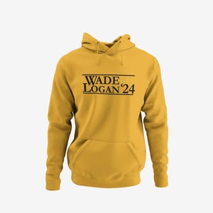 May include: Mustard yellow hoodie with a front pocket and drawstring hood. The hoodie features the text "WADE LOGAN '24" in black, centered on the chest. The garment is made of a soft material, suitable for casual wear.