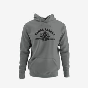 May include: A gray hooded sweatshirt with a front pocket and drawstring hood. The black graphic on the front reads "NANDA PARBAT" above "LEAGUE OF ASSASSINS" and "EST.1987", with a skull and crossed swords design.