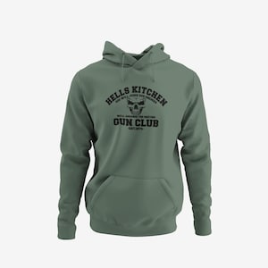 May include: Sage green hoodie with a front pocket and drawstring hood. The black graphic on the front reads "HELLS KITCHEN GUN CLUB" with a skull and the text "GOD WILL JUDGE OUR ENEMIES" and "WE'LL ARRANGE THE MEETING EST.1974."