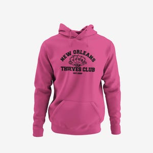 May include: A magenta hoodie with a front pocket and drawstring hood. The black graphic on the front reads "New Orleans Thieves Club" with playing cards and "Est. 1990."