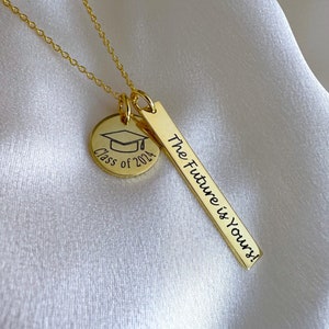 Puede incluir: Collar de oro con dos colgantes. El primer colgante es un disco redondo con el texto "Class of 2024" y un icono de gorro de graduación. El segundo colgante es una barra rectangular con el texto "The Future is Yours!"