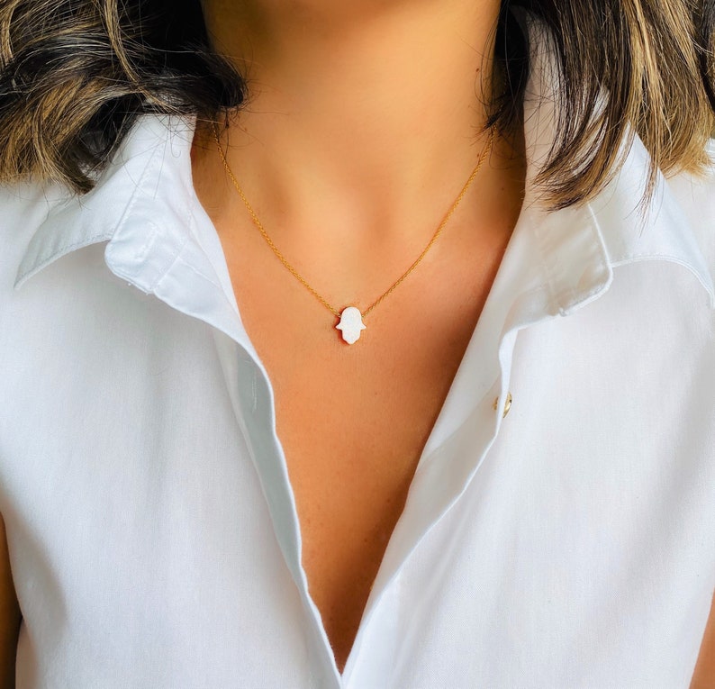 May include: A gold chain necklace with a white hamsa hand pendant.