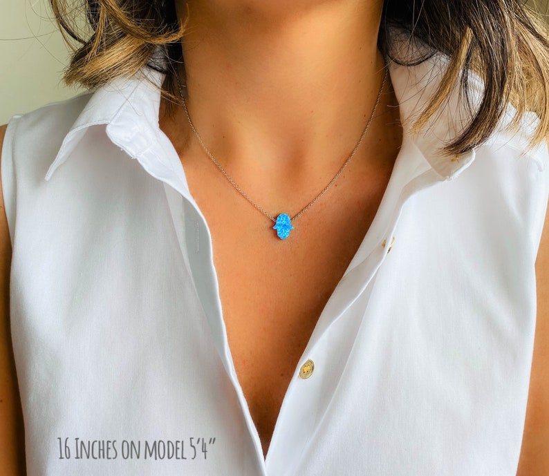 May include: A silver chain necklace with a blue opal hamsa hand pendant. The necklace is 16 inches long and is shown on a model. "16 inches on model 5'4" is written at the bottom of the image.