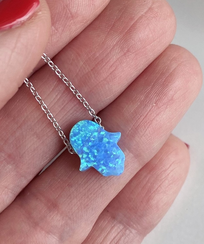 May include: A silver chain necklace with a blue opal hamsa hand charm. The hamsa hand is a symbol of protection and good luck.