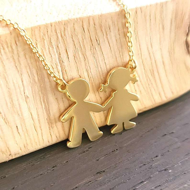 May include: Gold necklace with a pendant featuring two stylized figures of a boy and girl holding hands. The figures are cut out and have a shiny finish.