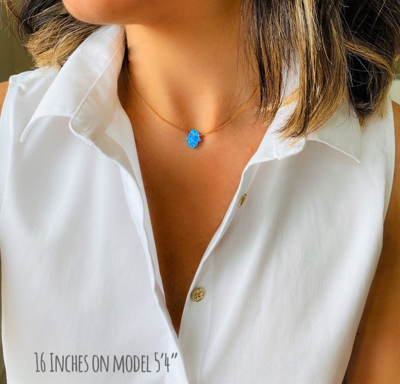 May include: A gold chain necklace with a blue opal hamsa hand charm. The necklace is 16 inches long and is shown on a model. "16 inches on model 5'4" is written at the bottom of the image.