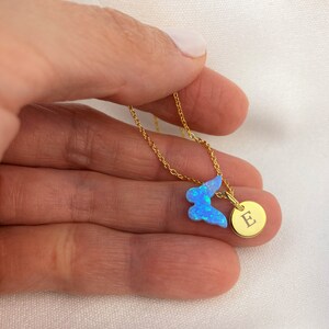 May include: A gold chain necklace with a blue opal butterfly charm and a gold disc charm with the letter "E" engraved on it.