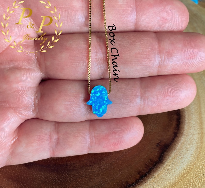 May include: A delicate gold chain necklace with a blue opal hamsa hand charm. The chain is a box chain style. The hamsa hand is a symbol of protection and good luck.