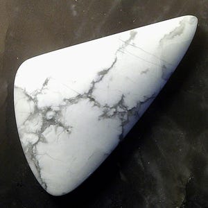 May include: A polished, triangular-shaped white stone with gray veining. The stone has a smooth, glossy surface and a rounded edge. The gray veining creates a marbled effect across the white surface. The stone is set against a dark background.