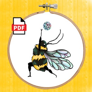 Puede incluir: Un patrón de punto de cruz digital en PDF que presenta un abejorro de dibujos animados en una pose dinámica, alcanzando una bola pixelada. El diseño está dentro de un bastidor de bordado, con un icono PDF en la esquina. La abeja tiene detalles en negro, amarillo y verde.