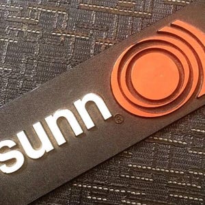 May include: A rectangular black sign with the white word "sunn" and an orange circular design. The sign has a textured surface and two small holes at the top. The design is a series of concentric circles.
