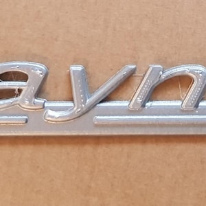Puede incluir: Un emblema de metal plateado con la palabra "Traynor" en relieve. El emblema tiene una línea horizontal elevada a cada lado de la palabra.