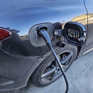 May include: A black electric car is plugged into a charging station. The charging cable is black and the charging port is on the driver's side rear fender. The car has a black wheel with silver spokes.