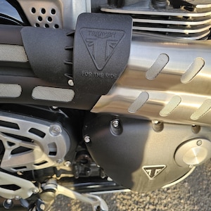 Könnte beinhalten: Nahaufnahme eines Motorradmotors mit einem schwarzen Hitzeschild, auf dem "Triumph" und "For the Ride" steht.