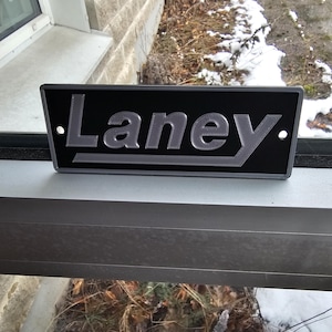 May include: A black metal sign with the word "Laney" in raised silver letters.
