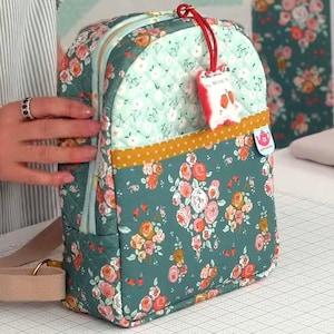 May include: A quilted backpack featuring a dark teal base with a floral pattern of pink and orange roses. The upper section is light mint green with a white grid pattern. A mustard yellow band separates the sections, and a small tag is attached.