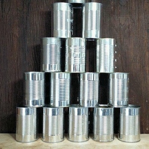 May include: A pyramid of silver metal cans, stacked against a dark wood background. The cans have ridged sides and are arranged in a triangular shape, with the base wider than the top.