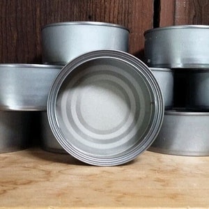 Könnte beinhalten: Eine Ansammlung leerer, silberfarbener Metallbehälter, die auf einer Holzoberfläche gestapelt und angeordnet sind. Die Behälter sind zylindrisch mit einem gerollten Rand und einem leicht vertieften Boden. Der Hintergrund ist eine dunkle Holzwand.