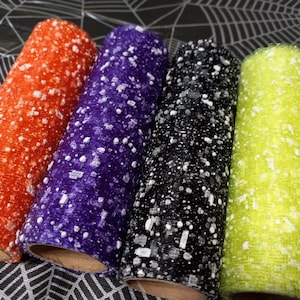 Puede incluir: Cuatro rollos de tela colorida, cada uno con un patrón moteado. Los colores incluyen naranja, morado, negro y verde lima. La tela está enrollada alrededor de un tubo de cartón. El fondo presenta un diseño de telaraña.