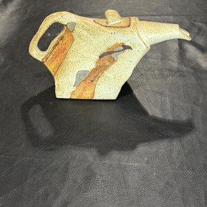 May include: A ceramic teapot with a unique, angular design. The teapot features a cream-colored base with abstract brown, orange, and blue accents. It has a curved handle and a long spout, perfect for serving tea.