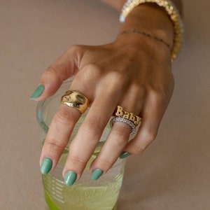 May include: A hand wearing three gold rings. The ring on the index finger is a thick, rounded band. The ring on the middle finger is a thin band with the word "Baby" spelled out in gold letters. The ring on the ring finger is a thin band with small clear stones. The hand is holding a glass of water with a lime wedge.