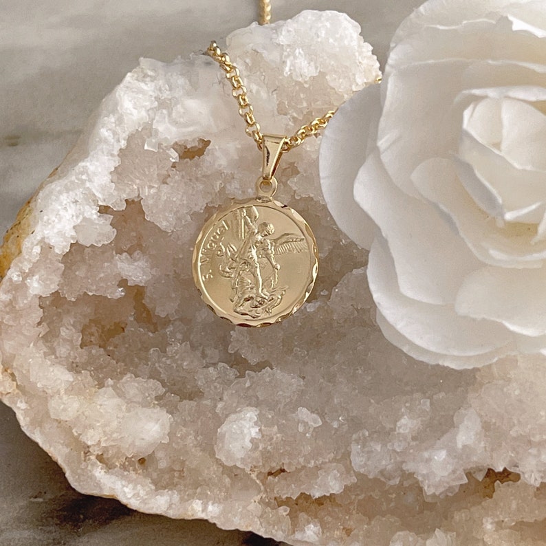 K&ouml;nnte beinhalten: Goldkette mit einem runden Anh&auml;nger, der ein detailliertes Bild eines Engels mit Fl&uuml;geln zeigt. Der Anh&auml;nger tr&auml;gt die Inschrift "St. Michael" am Rand.