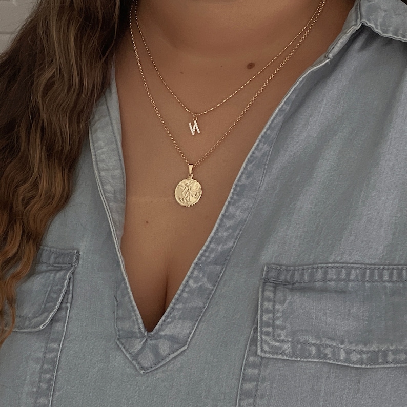 K&ouml;nnte beinhalten: Eine goldene Kettenkette mit einem kleinen M&uuml;nzenanh&auml;nger und einem Buchstaben 'N'-Anh&auml;nger. Die Kette ist &uuml;ber eine zweite goldene Kette gelegt.