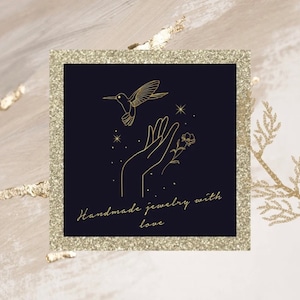 K&ouml;nnte beinhalten: Ein schwarzes Quadrat mit goldener Umrandung zeigt eine Linienzeichnung eines Kolibris, der zu einer offenen Hand mit einer Blume fliegt. Der Text "Handgemachte Schmuckst&uuml;cke mit Liebe" ist in goldener Schrift unter dem Bild geschrieben.
