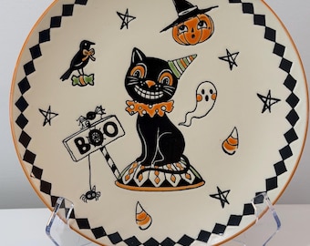 May include: A round ceramic plate with a cream-colored center and a black and white diamond-patterned border. The design features a black cat wearing a party hat and ruff, a pumpkin with a witch hat, a ghost, a crow, and a sign reading BOO.