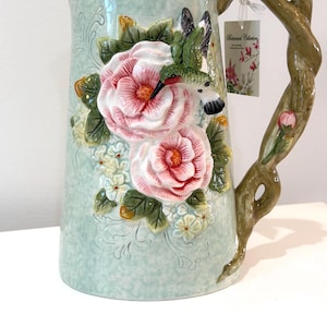 May include: A light blue ceramic pitcher with a detailed floral design. The pitcher features two large pink roses, white flowers, and a hummingbird. The handle is designed to look like a tree branch. A tag reads "Botanical Collection".