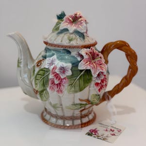 May include: A decorative ceramic teapot with a floral design. The teapot features pink and white flowers, green leaves, and a brown woven handle. The lid is adorned with a large pink flower. The teapot is on a white surface.