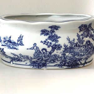 May include: A white ceramic planter with a blue and white pastoral scene. The oval-shaped planter has a scalloped rim and blue handles. The detailed design depicts figures, trees, and buildings in a classic toile style.