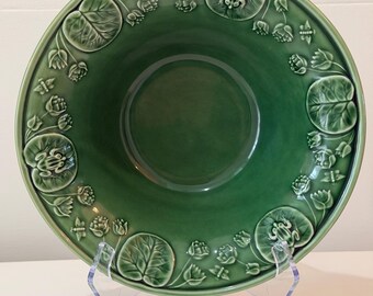 May include: A green ceramic serving bowl with an embossed design of lily pads, flowers, and frogs around the rim. The bowl is displayed on a clear acrylic stand. The bowl is a decorative piece.