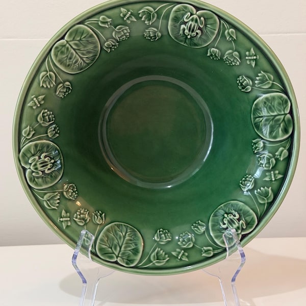 May include: A green ceramic serving bowl with an embossed design of lily pads, flowers, and frogs around the rim. The bowl is displayed on a clear acrylic stand. The bowl is a decorative piece.