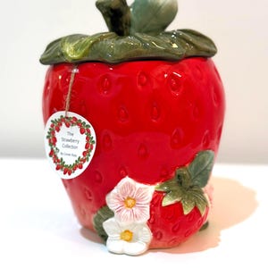 May include: A ceramic strawberry-shaped jar with a lid featuring green leaves and a stem. The jar is bright red with white flowers and a small strawberry accent. The tag reads "The Strawberry Collection".