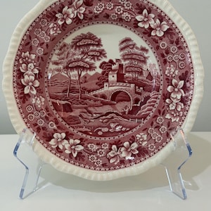 May include: A round ceramic plate with a scalloped edge featuring a deep red transferware pattern. The center depicts a landscape with a stone bridge, trees, and a building. The border is decorated with a repeating floral and botanical motif.