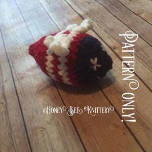 May include: A crocheted item in red, white, and navy blue, with a star detail. The item has a rounded shape and a textured appearance. The text "PATTERN ONLY!" and "HONEY BEE KNITTERY" are visible.