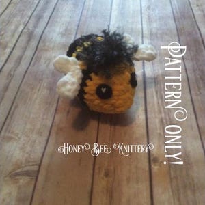 May include: A handmade crochet bee in yellow, black, and white. The bee has a fuzzy black head, white wings, and a small heart-shaped detail. The text "PATTERN ONLY!" and "HONEY BEE KNITTERY" are visible.