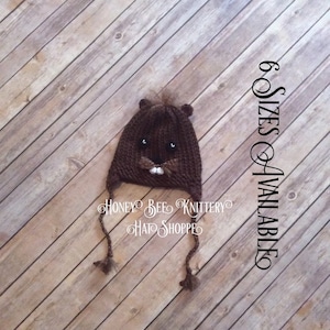May include: Brown knitted beaver hat with black eyes and white teeth. The hat has long ties that can be tied under the chin. The hat is on a wooden background. 6 Sizes Available.