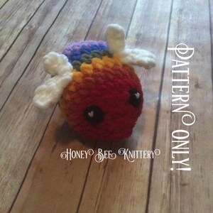 May include: A handmade crochet creature with a rainbow top and white wings. The body is red with black eyes and heart-shaped pupils. The text "PATTERN ONLY!" and "HONEY BEE KNITTERY" are visible.
